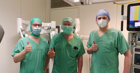 Dr. med. Yannick Lippka, Dr. med. Georg Schön und Dr. med. Marcel Stoll stehen in OP-Kleidung und recken den Daumen empor. 