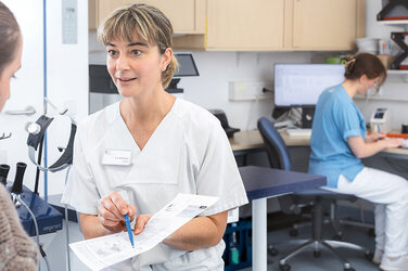 Ärztin Konstanze Beddermann zeigt einer Patientin einen Zettel und bereitet sie ihre OP vor. Hinter ihr sitzt die MFA Lena Supplieth und unterstützt sie 