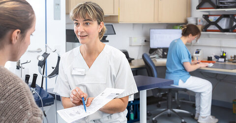 Ärztin Konstanze Beddermann zeigt einer Patientin einen Zettel und bereitet sie ihre OP vor. Hinter ihr sitzt die MFA Lena Supplieth und unterstützt sie 