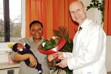 Prof. Bader überreicht Edwige Tiepokin, die ihre Tochter Christi im Arm hält, einen Blumenstrauß.