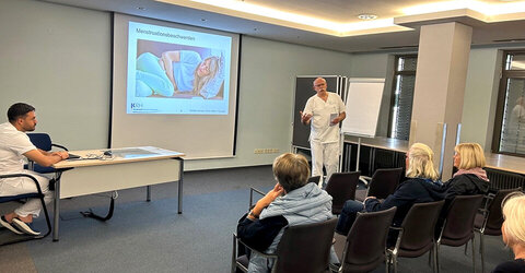 Chefarzt Dr. med. Hubert K. Sommer steht vor einer Leinwand und trägt vor. Vor ihm sitzen interessierte Frauen
