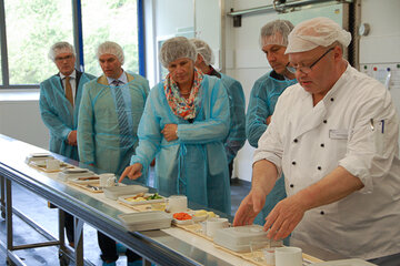 Wolfgang Faustin erklärt den Besuchern am Portionierungsband, wie das Essen auf die Teller  und Tabletts gelangt.